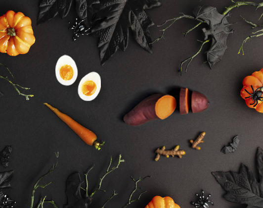 Various Halloween-themed decorations arranged on a dark background, including miniature orange pumpkins, black leaves, rubber bats and spiders, alongside food items such as a sliced sweet potato, a whole carrot, two halved boiled eggs and small pieces of ginger.